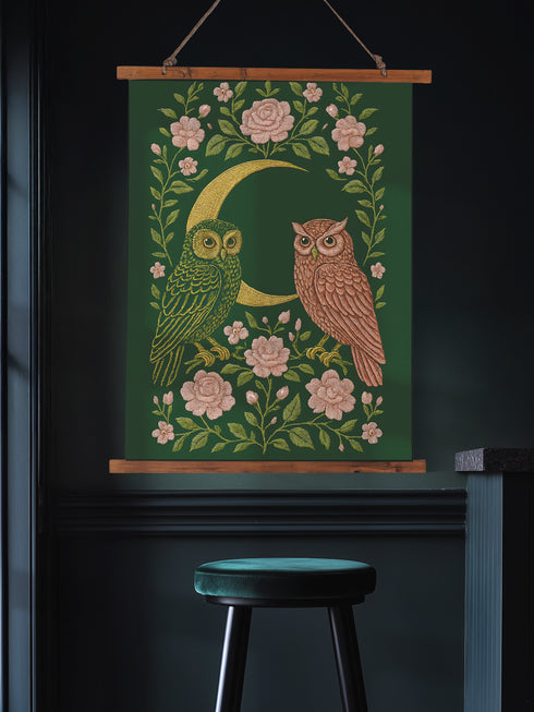 26x36 inch vertical wall tapestry printed on silky polyester, featuring a pair of embroidered-style owls perched on a golden crescent moon, surrounded by pink vintage-style florals and greenery on a forest green background. Includes stained wood slats and twine for easy hanging. A romantic, nature-inspired design for woodland and cottagecore decor lovers.